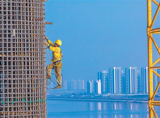 奮戰(zhàn)在百米索塔之上的建設(shè)者身影，與遠處的高樓大廈遙呼相應(yīng)。